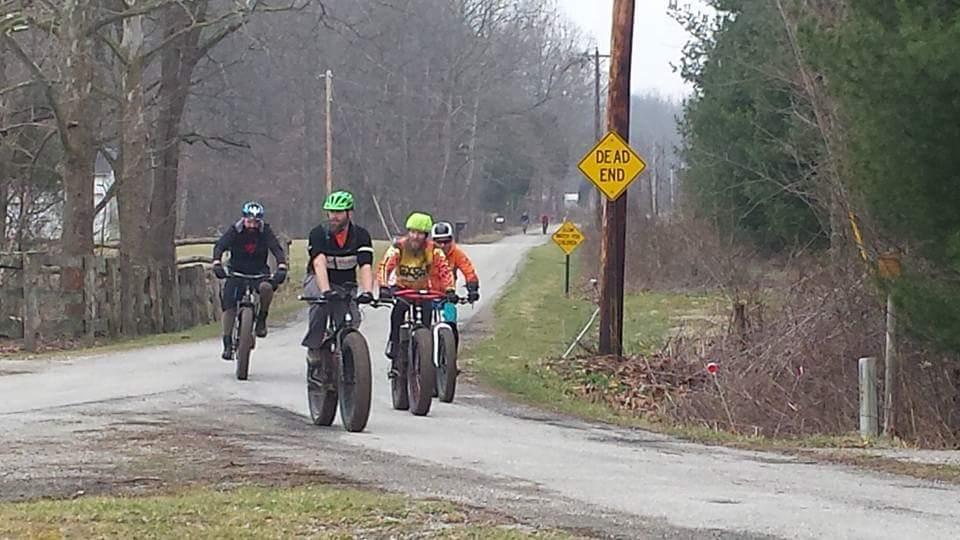 Four cyclists riding fat tire bikes on a rural road, with a "Dead End" road sign visible. The scene features trees lining both sides of the road and a cloudy sky in the background. Versailles State Park mountain bike trail.