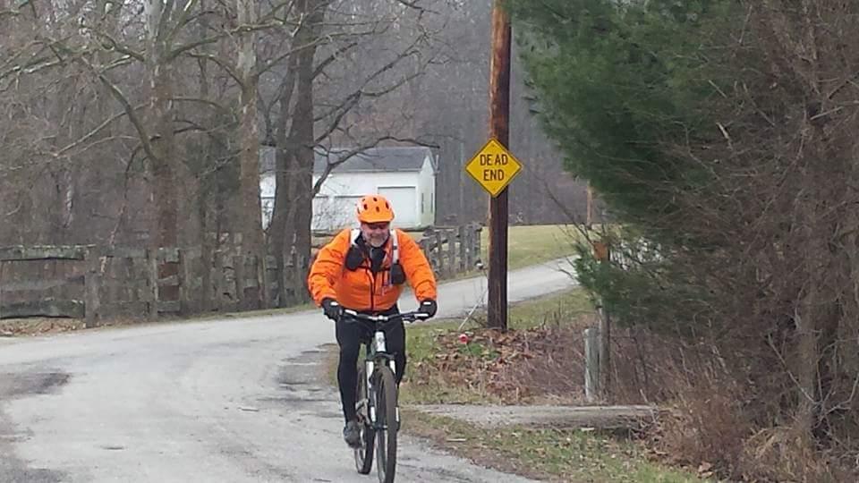 A person biking on a rural road in cloudy weather, wearing an orange jacket and helmet, with a "Dead End" sign visible nearby and a wooden fence along the path. Trees line the road, and a house is seen in the background. Versailles State Park mountain bike trail.