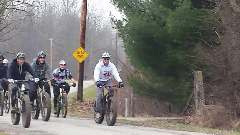 A group of six cyclists riding on a rural road, with a "Dead End" sign in the background. The cyclists are wearing various types of athletic clothing and helmets, and are riding mountain bikes. Trees and a grassy area are visible alongside the road, suggesting an outdoor scene in a wooded environment. Versailles State Park mountain bike trail.