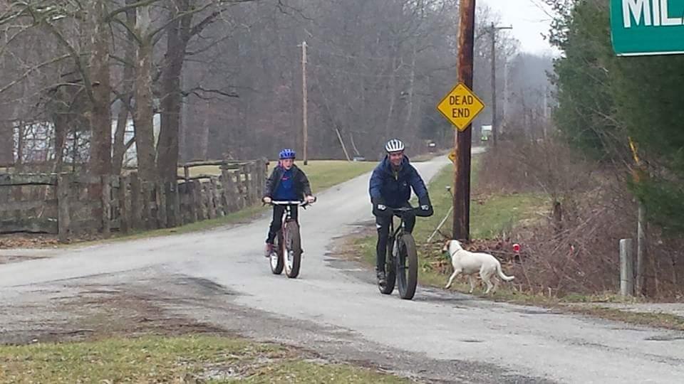 Two individuals riding bicycles along a rural road with a "Dead End" sign visible. One cyclist is on a fat bike, while the other rides a standard bike. A white dog is walking alongside the cyclists on the roadside, and there are trees and a wooden fence in the background. Versailles State Park mountain bike trail.