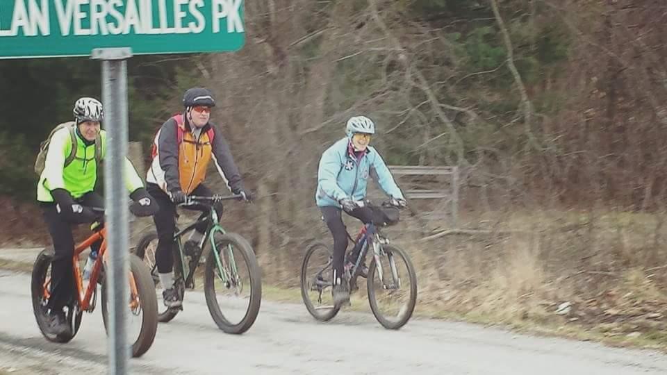 Image of three cyclists riding on a gravel path near a road sign indicating "VERSAILLES PK." They are wearing various cycling attire, including bright colors for visibility. The surrounding landscape features bare trees and a natural setting, suggesting an outdoor recreational activity. Versailles State Park mountain bike trail.