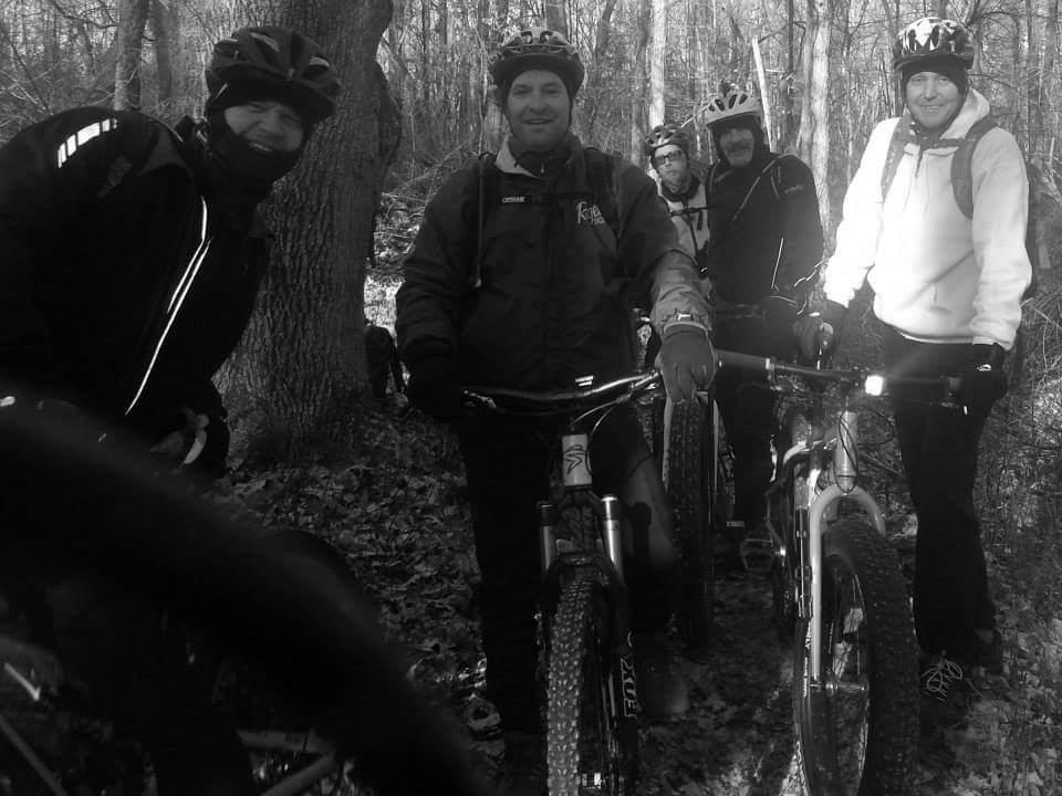 A group of five mountain bikers posing for a photo in a wooded area, surrounded by trees and fallen leaves. They are wearing winter biking gear, including helmets and jackets, and are standing next to their bikes. The image is in black and white, capturing the camaraderie of the group during their ride. Versailles State Park mountain bike trail.