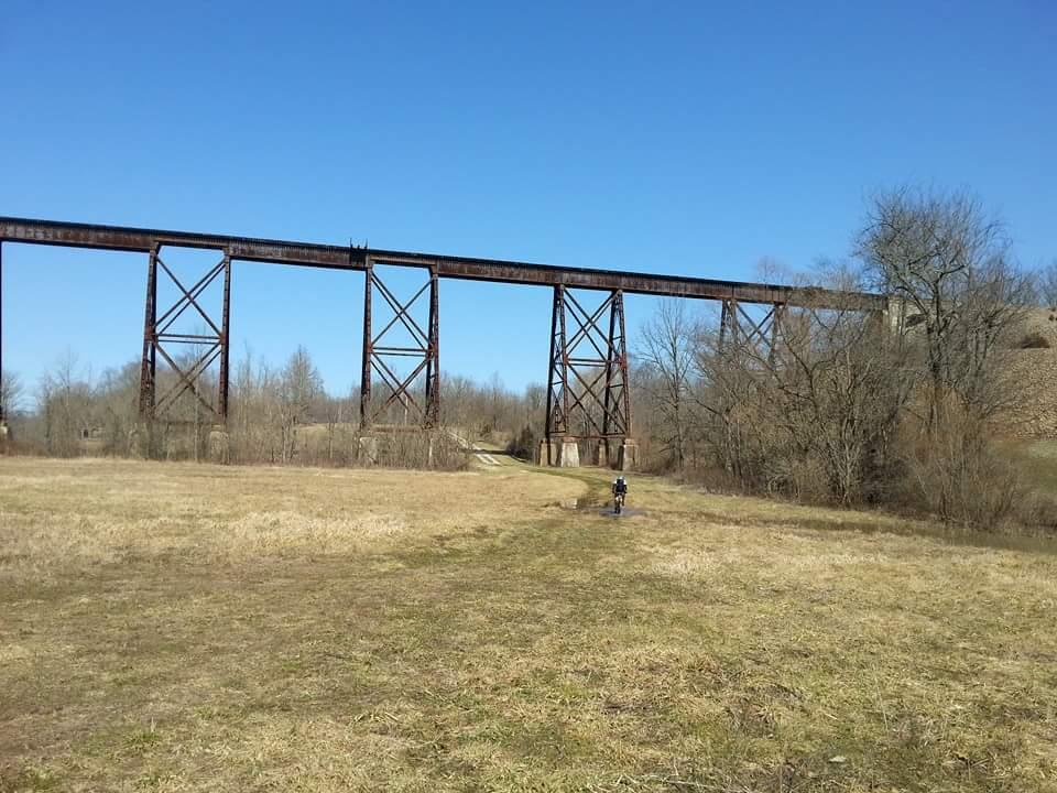 A large, rusty rail bridge towers above a grassy field, under a clear blue sky. A person is walking along a path in the foreground, with trees and brush scattered on either side. The bridge's towering structure and the serene landscape create a contrast between the man-made and natural environments. Versailles State Park mountain bike trail.