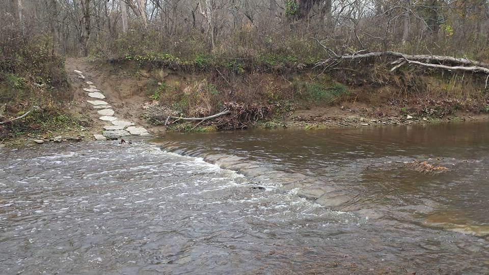 A tranquil scene of a shallow river with gently flowing water, bordered by a rocky stream bed. On the left, a stone pathway leads up to higher ground, surrounded by trees and sparse shrubbery. The area appears to be in a natural setting with muted colors, suggesting an overcast day. Versailles State Park mountain bike trail.