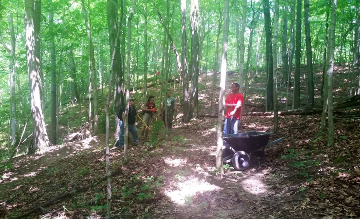 A group of people engaged in outdoor work in a green, forested area. Some individuals are holding tools, while one person stands next to a wheelbarrow on a leaf-covered trail. Sunlight filters through the trees, creating a dappled effect on the ground. The scene depicts community involvement in a nature conservation or trail maintenance activity. Versailles State Park mountain bike trail.