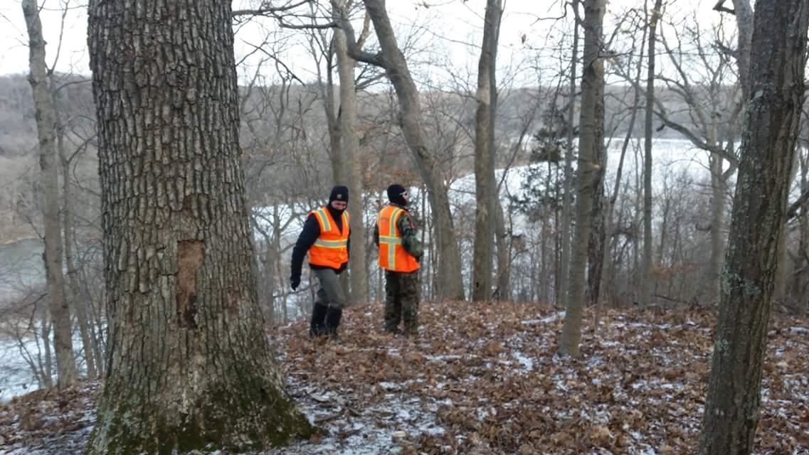 Two individuals wearing bright orange safety vests and winter clothing walk through a forested area with bare trees and a light dusting of snow on the ground. A river is visible in the background, reflecting a winter landscape. Versailles State Park mountain bike trail.