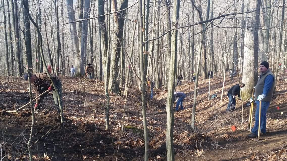 A group of volunteers planting trees or clearing land in a forested area during the winter months. The scene includes several people using shovels and other tools among bare trees and fallen leaves, with an emphasis on environmental conservation efforts. Versailles State Park mountain bike trail.