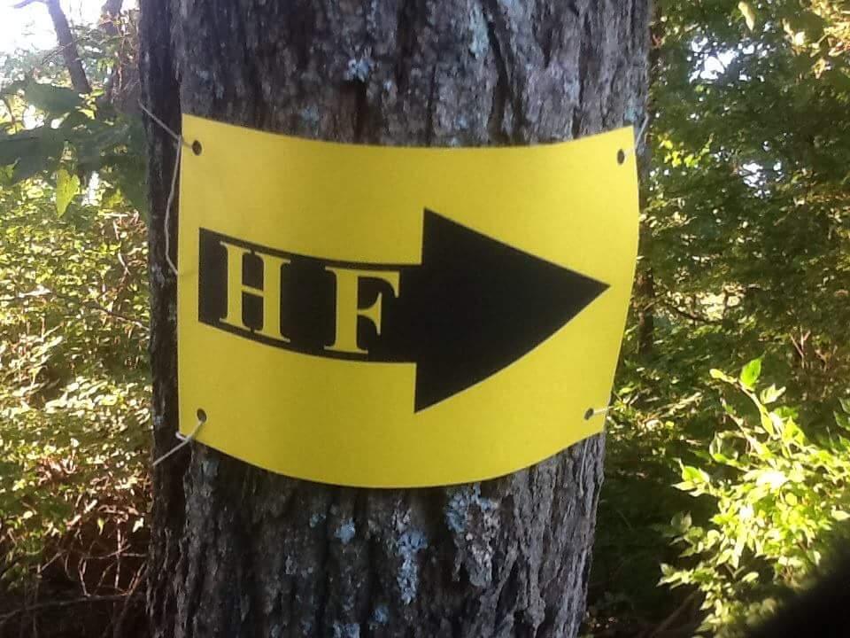 A yellow sign affixed to a tree, featuring the letters "HF" in bold black text and a large black arrow pointing to the right. Surrounding foliage is visible, indicating a natural outdoor setting. Versailles State Park mountain bike trail.