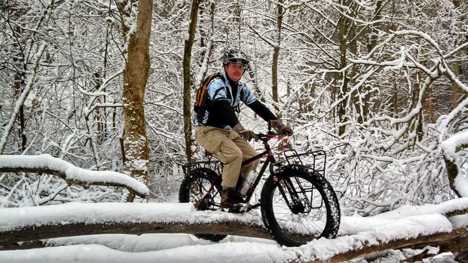 A person riding a mountain bike on a snow-covered fallen log in a winter forest. Surrounding trees are blanketed in snow, creating a serene and picturesque winter landscape. The cyclist is wearing a helmet and winter gear, highlighting the adventurous spirit of biking in snowy conditions. Versailles State Park mountain bike trail.