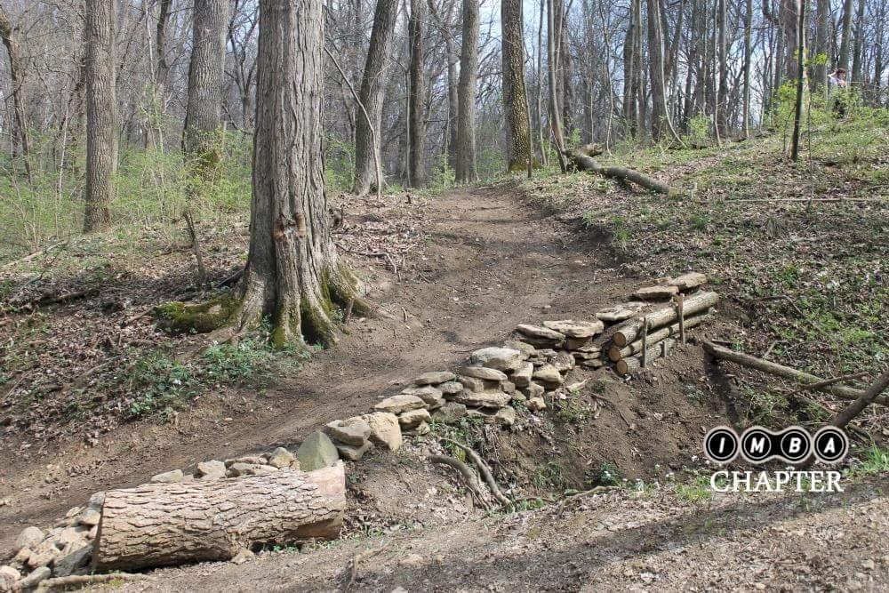 A dirt pathway through a wooded area, featuring a stone and log structure built into the trail. There are tall trees and sparse underbrush surrounding the area, with early spring foliage visible. The image appears to be taken in a forest setting, highlighting trail maintenance efforts. Harbin park mountain bike trail.