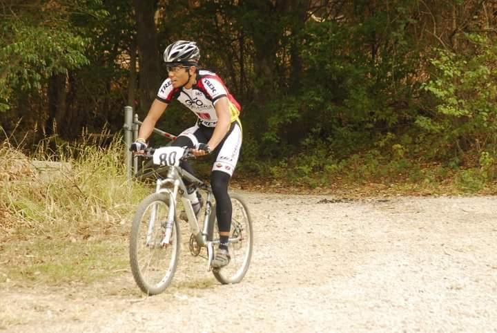 A cyclist wearing a racing outfit and helmet is riding a mountain bike on a gravel path surrounded by trees. The rider has a race number 80 visible on their jersey. Tower Park mountain bike trail.