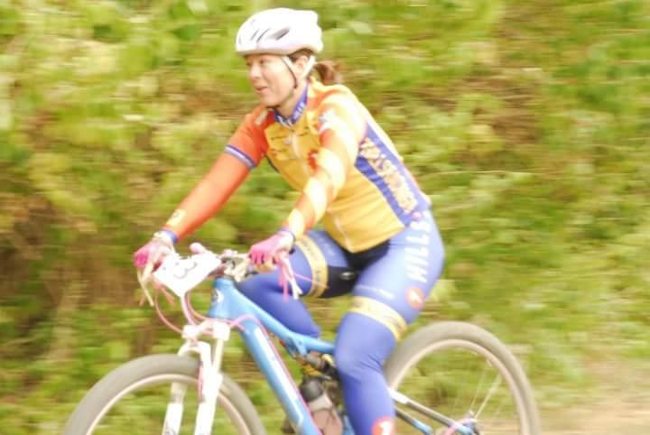A female cyclist wearing a brightly colored jersey and helmet rides a mountain bike on a trail surrounded by greenery. The image is slightly blurred, capturing the motion of the ride. Tower Park mountain bike trail.