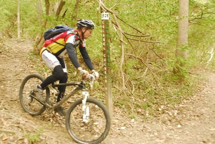 A mountain biker riding along a dirt trail surrounded by greenery, with a trail marker post indicating the trail number in the background. The cyclist is wearing a helmet and colorful biking gear, and the terrain appears to be natural and uneven. Tower Park mountain bike trail.
