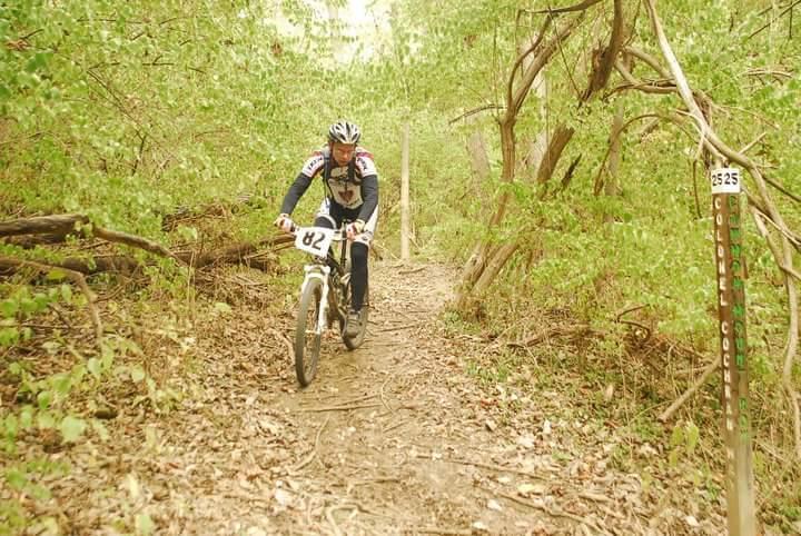 A mountain biker wearing a helmet and jersey navigates a narrow, leafy path in a wooded area. The trail is marked with a sign indicating "Colonel Cochrane" and is surrounded by vibrant green foliage and fallen leaves. Tower Park mountain bike trail.