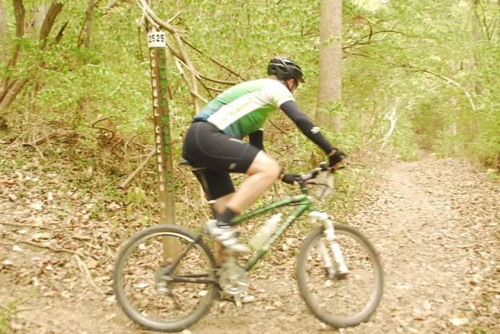 A mountain biker in a green and white jersey rides along a dirt trail surrounded by trees. A trail marker with the number 625 is visible nearby, indicating the path. The ground is covered with fallen leaves, and the scene conveys a sense of nature and outdoor adventure. Tower Park mountain bike trail.
