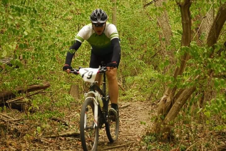 A cyclist wearing a helmet and a green and white jersey rides a mountain bike on a narrow trail surrounded by dense greenery. The cyclist has a race number, 79, displayed on the front of their bike. Tower Park mountain bike trail.