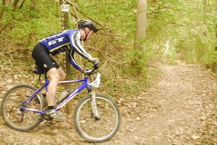 A cyclist in a blue and black jersey rides a mountain bike along a dirt trail surrounded by leafy trees, navigating a turn. The scene is set in a natural, wooded area with fallen leaves on the ground, suggesting an outdoor biking adventure. Tower Park mountain bike trail.