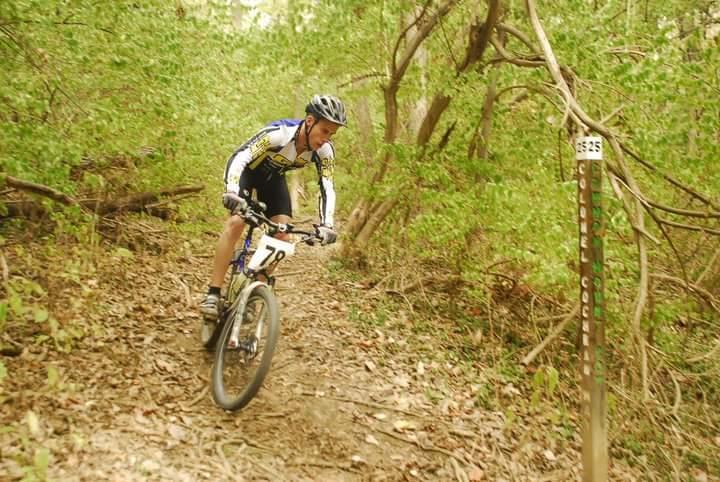 A mountain biker wearing a helmet and cycling gear navigates a narrow, dirt trail surrounded by greenery, with a trail marker visible on the right side of the image. Tower Park mountain bike trail.