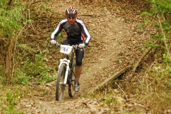 A mountain biker wearing a helmet and racing gear navigates a dirt trail surrounded by trees and foliage. The cyclist is focused and in motion, with the number "44" displayed on his jersey. The ground is covered with fallen leaves and there are some rocky sections along the path. Tower Park mountain bike trail.