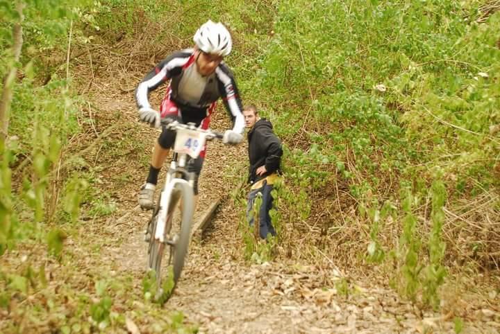 A mountain biker in a black and red jersey rides along a dirt trail surrounded by greenery, while a second person in a black jacket watches from the side of the trail. Tower Park mountain bike trail.