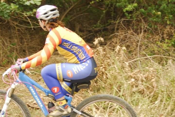 A cyclist wearing a colorful jersey and helmet rides a mountain bike along a dirt trail surrounded by greenery and dried foliage. Tower Park mountain bike trail.