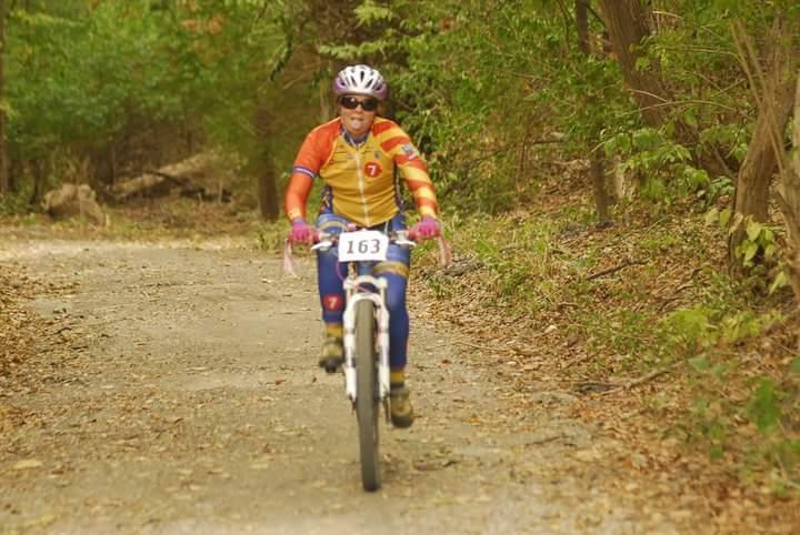 A cyclist in colorful biking gear rides on a gravel path surrounded by trees. The rider, wearing a helmet and sunglasses, has a race number pinned to their outfit and appears to be enjoying the ride. The scenery includes fallen leaves and lush greenery typical of a forest environment. Tower Park mountain bike trail.