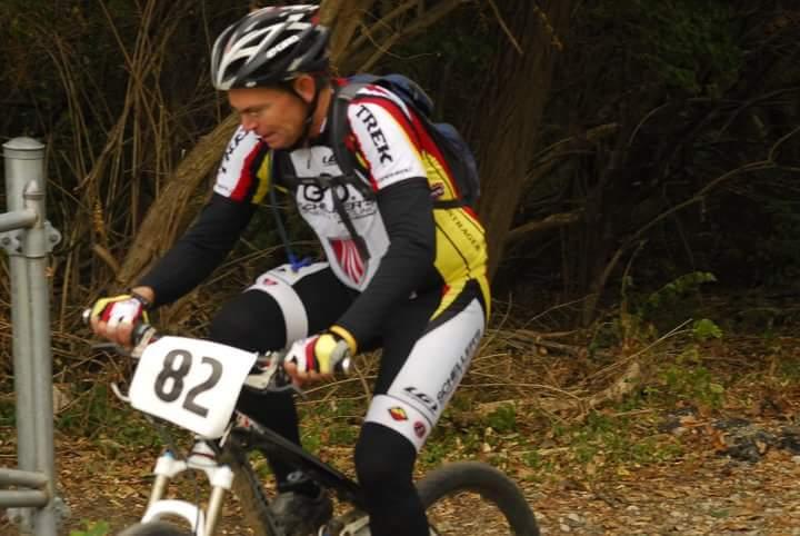 A cyclist wearing a racing helmet and a colorful biking jersey, featuring the number 82, rides along a gravel path surrounded by trees and natural foliage. The cyclist is focused on navigating the terrain, showcasing an active outdoor environment. Tower Park mountain bike trail.
