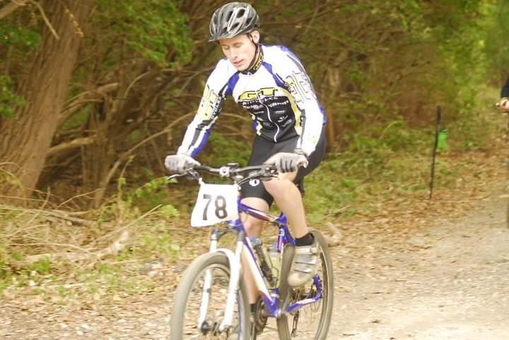 A mountain biker wearing a white and blue jersey with the number 78 rides along a dirt path surrounded by trees. The cyclist is focused on navigating the terrain, wearing a helmet and gloves for safety. Tower Park mountain bike trail.