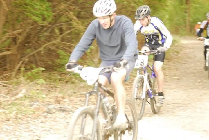 Two mountain bikers riding on a wooded dirt trail. One rider is wearing a gray sweatshirt and a white helmet, while the other is in a colorful cycling jersey and black helmet. The image captures a sense of motion, with both cyclists pedaling actively. Tower Park mountain bike trail.
