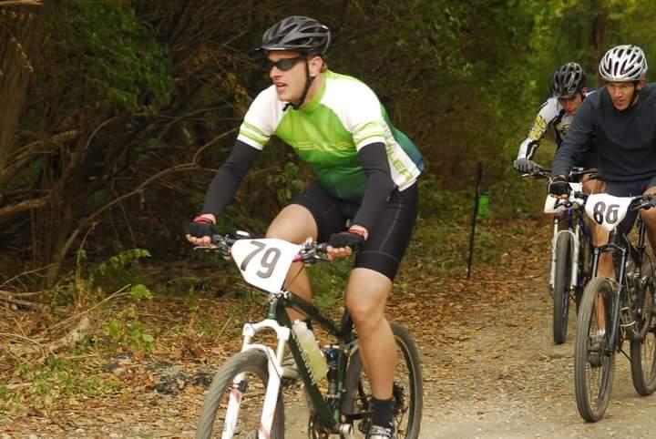 A group of three cyclists competes in a mountain biking event on a gravel trail surrounded by trees. The rider in the foreground, wearing a green and black jersey with number 79, is focused on navigating the trail. Two other cyclists can be seen behind him, one in a dark shirt and the other in a white jersey with number 86. The scene is set in a natural outdoor environment, suggesting an active and competitive atmosphere. Tower Park mountain bike trail.