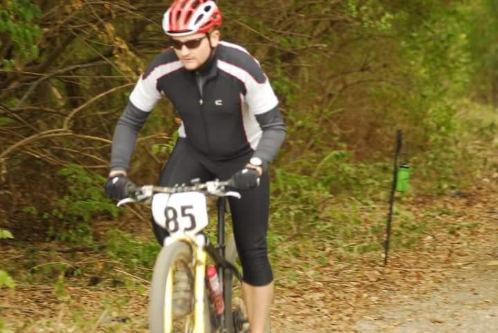 A cyclist in a black and white racing jersey and red helmet rides a mountain bike along a gravel trail surrounded by greenery. The bike has a number "85" displayed on the front. Tower Park mountain bike trail.