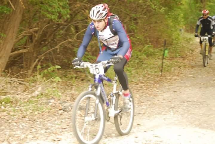 A cyclist in a blue and red racing outfit is riding a mountain bike along a dirt trail surrounded by greenery. The rider has a helmet and a race number visible on their chest. In the background, another cyclist can be seen riding in a black outfit. The scene depicts an outdoor cycling event. Tower Park mountain bike trail.