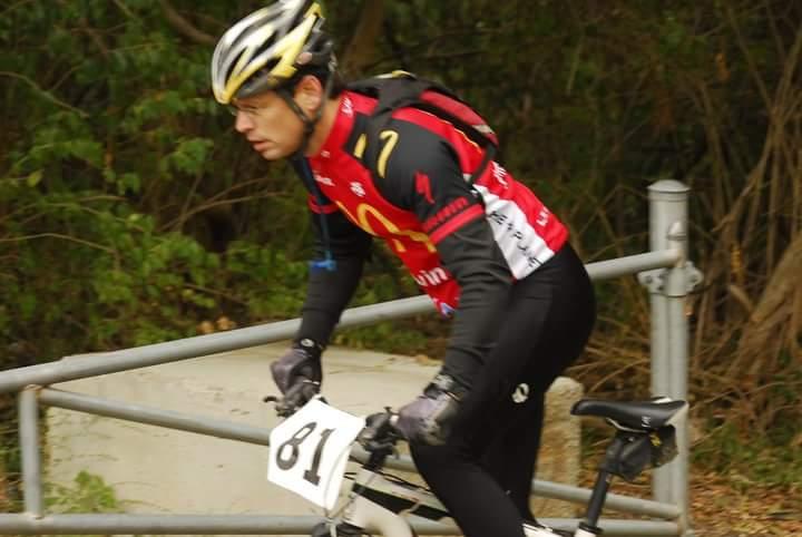A cyclist in a black and red racing suit, wearing a helmet, focuses intently while riding a mountain bike. The bike has a race number, 81, attached to it. The background features greenery and a fence, suggesting an outdoor racing event. Tower Park mountain bike trail.
