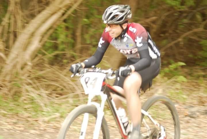 A cyclist in a black and red jersey rides a mountain bike on a dirt trail, surrounded by trees. The image captures the motion and speed of the athlete. The cyclist's number is visible on the bike, indicating participation in a race or event. Tower Park mountain bike trail.