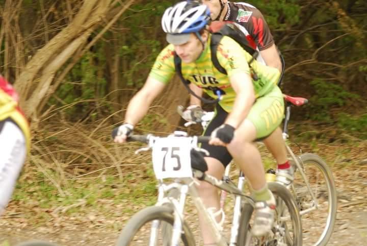 A group of mountain bikers navigating a dirt trail in a wooded area. One cyclist, wearing a green and yellow jersey with the number 75, is in the foreground, focused on the path ahead. Another cyclist, dressed in a black and red jersey, is following closely behind. The background features trees and foliage, indicating a natural outdoor setting. Tower Park mountain bike trail.