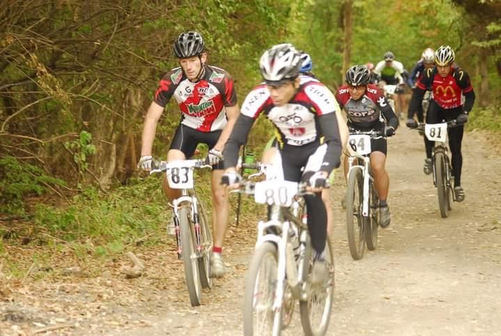 A group of male mountain bikers, wearing racing jerseys and helmets, navigates a gravel trail surrounded by trees. The cyclists are numbered from 80 to 84, with one cyclist in the foreground wearing a red and black jersey. The scene captures a competitive and energetic atmosphere during an outdoor race. Tower Park mountain bike trail.