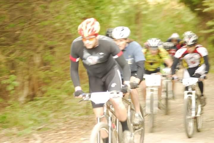 A group of mountain bikers racing on a wooded trail. The image shows several cyclists in motion, some wearing helmets and racing gear, as they navigate the path surrounded by greenery. The photo has a slight blur effect, emphasizing the speed of the riders. Tower Park mountain bike trail.
