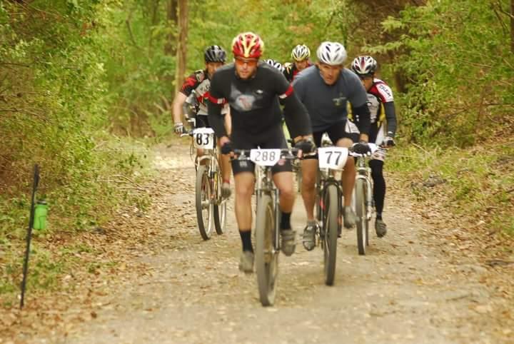 A group of mountain bikers navigating a dirt trail surrounded by trees, with several cyclists in the foreground focused on their race. The riders are wearing helmets and numbered jerseys, showcasing their competitive spirit in an outdoor event. Tower Park mountain bike trail.