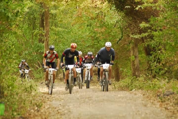 A group of mountain bikers riding along a dirt trail in a forested area, surrounded by greenery. The cyclists are wearing helmets and race numbers, showing a mix of focused expressions as they navigate the rugged terrain. Tower Park mountain bike trail.