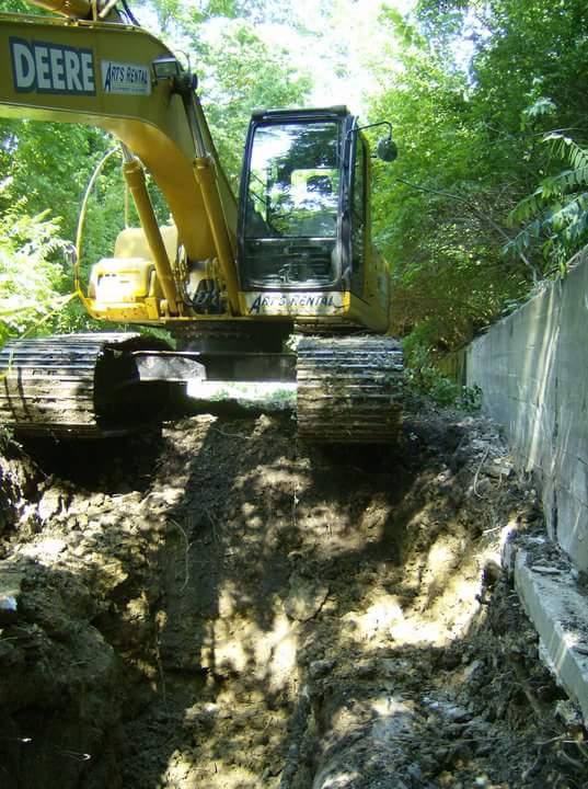 A yellow excavator positioned on a mound of dirt, surrounded by greenery. The machine is facing a vertical concrete wall, with deep trenches visible in the soil below. Sunlight filters through the trees, illuminating the scene. Tower Park mountain bike trail.