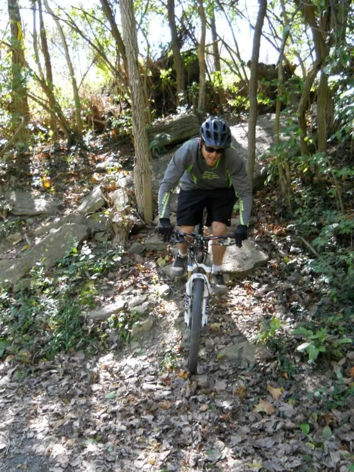 A mountain biker navigates a rocky and leafy trail surrounded by trees, wearing a helmet and sunglasses, showcasing determination as he descends the path. Tower Park mountain bike trail.