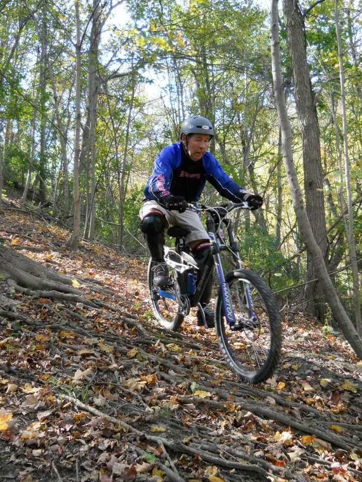 A mountain biker navigating a sloped trail covered in autumn leaves and tree roots, surrounded by lush green trees in a forested area. The rider wears a helmet and protective gear, showcasing focused concentration while descending the path. Tower Park mountain bike trail.