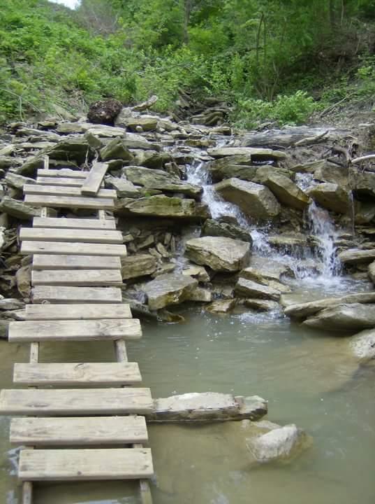 A wooden bridge made of planks crosses over a small stream with cascading water, surrounded by rocky terrain and lush greenery. The scene depicts a serene outdoor setting, highlighting nature's tranquility. Tower Park mountain bike trail.