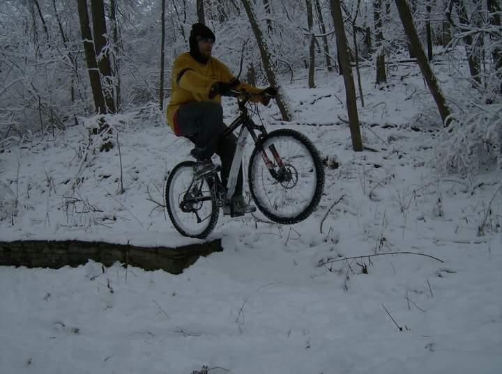 A person wearing a yellow hoodie and grey pants is riding a mountain bike, jumping over a small ledge in a snowy forest. Snow-covered trees surround them, creating a winter atmosphere. Tower Park mountain bike trail.