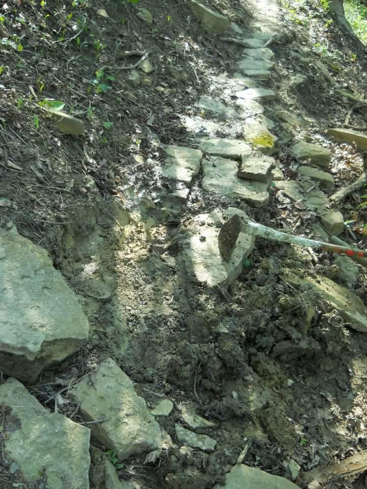 A partially completed stone pathway on a dirt hillside, with some loose rocks and a shovel resting on the ground. The area appears to be in a forested setting, with green vegetation visible. Tower Park mountain bike trail.