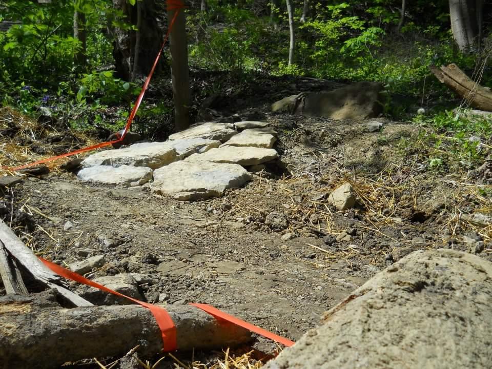 A partially constructed pathway in a lush green forest, featuring large stones arranged on a dirt surface. There are orange construction ribbons marking the area, along with scattered debris like fallen branches and earthy materials. Tower Park mountain bike trail.