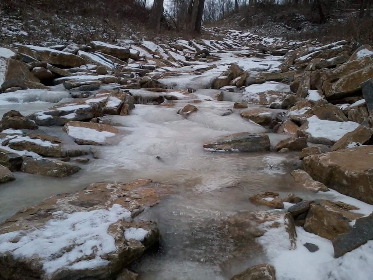 A frozen stream surrounded by large rocks, with patches of snow visible. The scene captures a cold, wintry landscape with water flowing between the stones, highlighting the textures of the ice and the rugged terrain. Tower Park mountain bike trail.