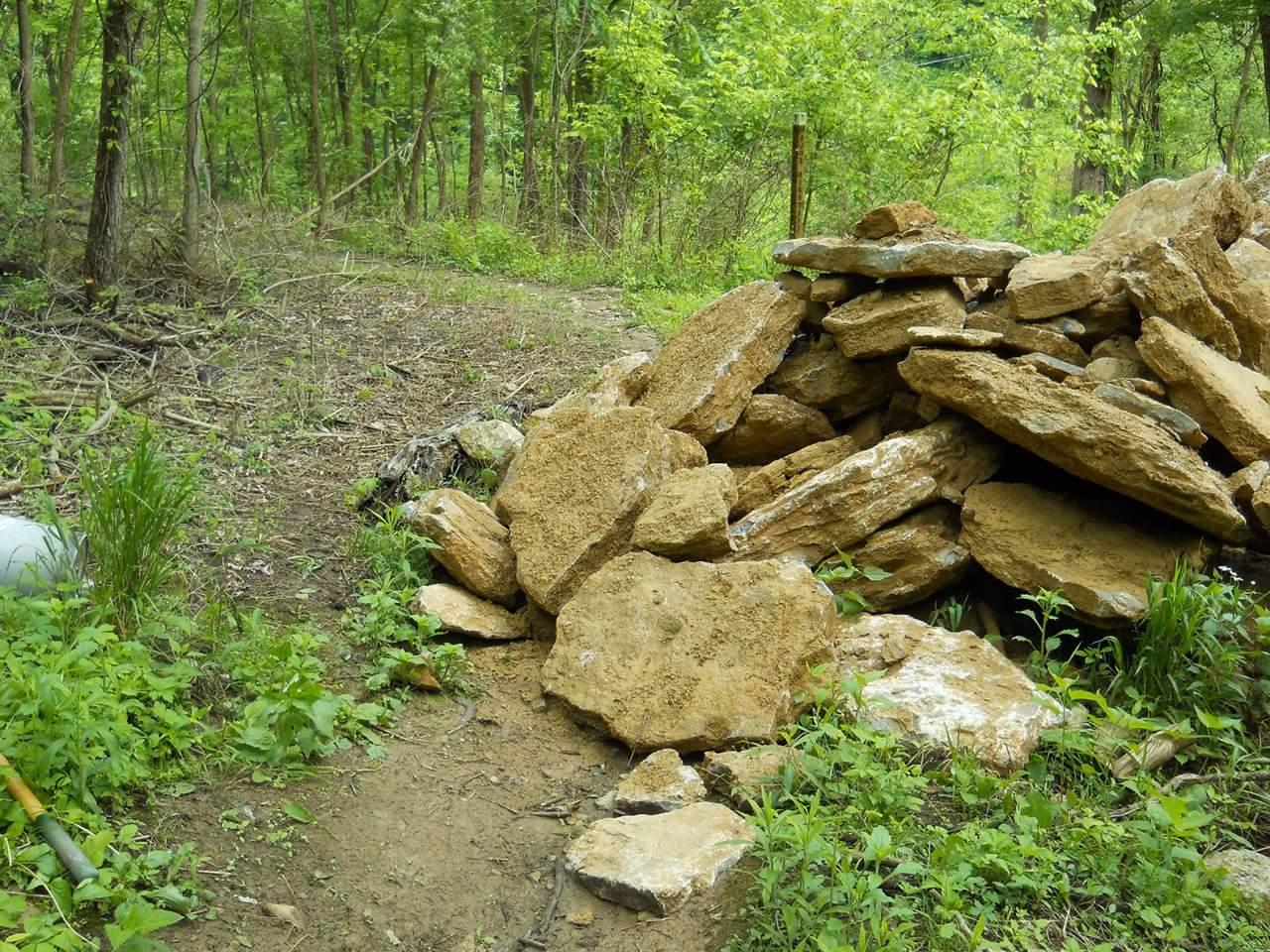 A pile of large, irregularly shaped rocks sits at the edge of a dirt path surrounded by greenery in a forested area. Lush vegetation is visible on both sides of the path, indicating a natural setting. Tower Park mountain bike trail.