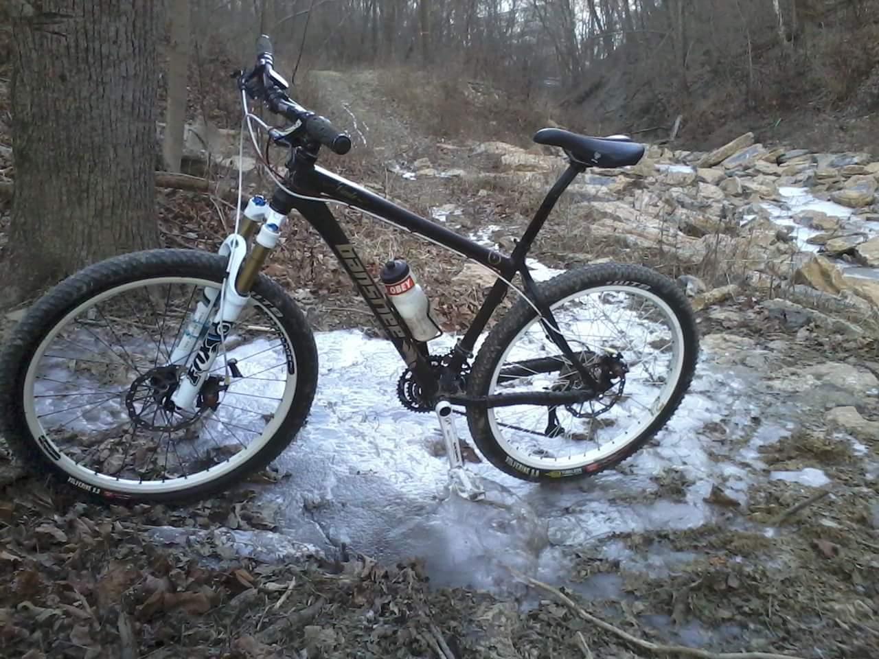 A black mountain bike parked beside a rocky stream in a wooded area. The bike has prominent suspension components, large tires, and a water bottle attached. Ice and water cover part of the ground near the bike, indicating a cold environment. Bare trees and dry leaves surround the scene. Tower Park mountain bike trail.