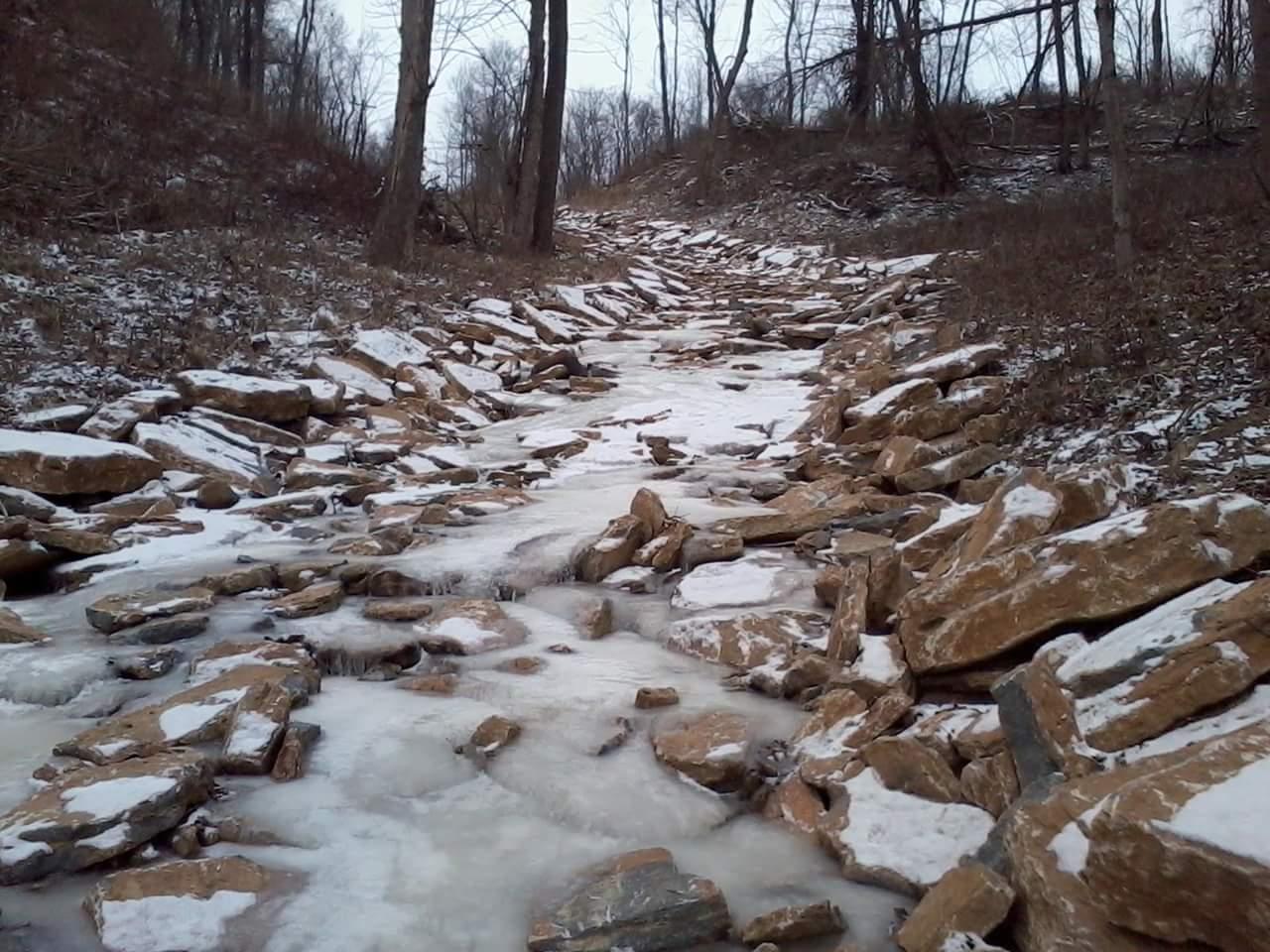 A rocky creek bed partially covered in ice and snow, surrounded by bare trees and winter foliage. The water is mostly frozen, with some flowing areas visible among the stones. Tower Park mountain bike trail.